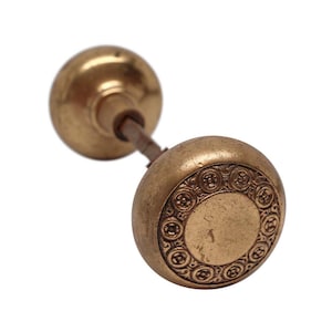 May include: Antique brass doorknob with a decorative floral pattern around the edge of the larger knob. The doorknob has a round shape and a tarnished finish, suggesting age and use. The smaller knob is also round.