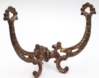 Può includere: Gancio da parete in metallo in stile antico e ornato con finitura bronzo. Il gancio presenta un design curvo con dettagli decorativi floreali e volute. Le estremità hanno un design circolare con un foro. Il gancio è progettato per essere montato a parete.