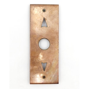 May include: A rectangular, copper-toned elevator button panel. It has an upward-pointing arrow, a circular button, and a downward-pointing arrow. The panel shows signs of age with visible wear and tear.