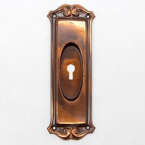 May include: A vintage, ornate, copper-colored keyhole cover with a decorative design. The cover has a rectangular shape with an oval keyhole cutout.