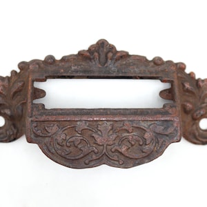 May include: A dark brown, rusted metal drawer pull with a rectangular label holder slot in the centre. The hardware features an ornate, raised floral and leaf relief pattern across the front surface and includes two circular screw holes on sides.