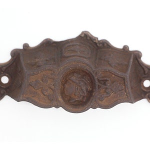 May include: A cast iron drawer pull with an ornate design. The pull features a central oval with a woman's face in profile, surrounded by floral motifs. The pull is a dark brown color and shows signs of age and wear.