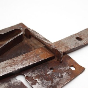 May include: Close-up of a rusty, antique metal latch. The latch features a rectangular base with a curved handle, a long, flat bar, and a textured, cylindrical handle. The metal has a weathered, aged appearance.