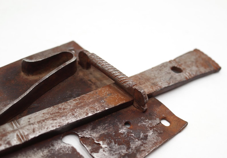 May include: Close-up of a rusty, antique metal latch. The latch features a rectangular base with a curved handle, a long, flat bar, and a textured, cylindrical handle. The metal has a weathered, aged appearance.