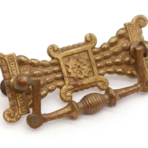 May include: A brass drawer pull with a decorative design featuring a beaded pattern and a central square with a floral motif. The pull has a curved handle.