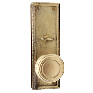 Puede incluir: Un juego de manija de puerta de latón de estilo vintage. El juego incluye una placa trasera rectangular con un ojo de cerradura y un pomo redondo. El acabado tiene una apariencia envejecida, lo que sugiere una estética clásica o antigua.