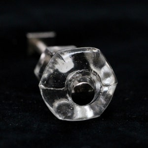 Puede incluir: Un tapón de botella de vidrio transparente con forma hexagonal y un tapón de rosca de metal plateado.