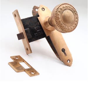 Puede incluir: Juego de herrajes de puerta de latón antiguo, que incluye un pomo redondo detallado, una placa para la cerradura y un mecanismo de pestillo rectangular. El conjunto tiene una estética vintage, con desgaste visible.