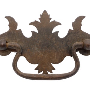 May include: A rustic, antique-style metal drawer pull with a decorative, ornate design. The pull has a curved handle and a central, decorative plate with a floral motif.