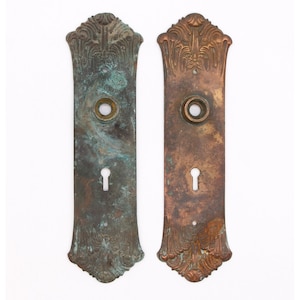 May include: Two antique door plates with ornate designs. The plate on the left is a greenish-brown colour, while the plate on the right is a more reddish-brown colour.