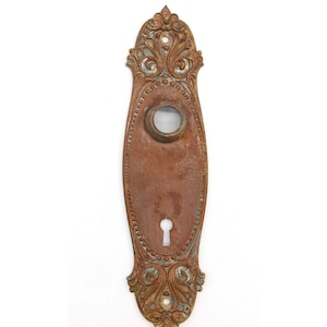 May include: Antique brass door plate with ornate detailing and a keyhole. The plate has a worn, aged patina.