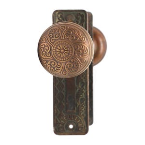 Może przedstawiać: Antyczny mosiężny klamka do drzwi z ozdobnym wzorem. Klamka jest przymocowana do mosiężnej płytki z ozdobnym wzorem.