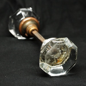 Pode incluir: Um puxador de porta de vidro transparente e facetado com uma haste de latão e bronze. O puxador tem uma estética vintage e está sobre um fundo escuro. O vidro está ligeiramente turvo, sugerindo idade.