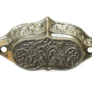 May include: Antique metal drawer pull with ornate floral and scrollwork design. The hardware has a distressed, aged appearance with visible rust and two mounting holes. The decorative piece is a dark gray color.
