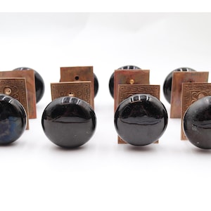 Może przedstawiać: Zestaw ośmiu czarnych ceramicznych gałek do szafek z błyszczącym wykończeniem. Każda gałka połączona jest z kwadratową, miedzianą płytką tylną z dekoracyjnym geometrycznym wzorem. Idealne do renowacji mebli.