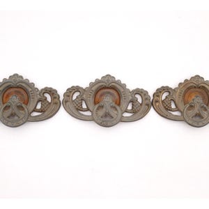 May include: Three antique bronze drawer pulls with ornate detailing. Each pull features a central ring handle, surrounded by decorative elements and a circular wooden accent. The pulls are arranged horizontally against a white background.
