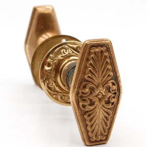 May include: Antique brass door hardware set. The set includes a decorative, elongated, octagonal door knob with an ornate floral design. The set also includes a round backplate with an embossed floral pattern. The hardware is set against a white background.