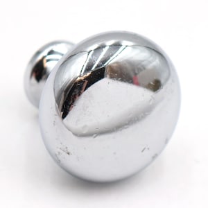 May include: A polished chrome cabinet knob with a smooth, rounded, mushroom-shaped head and a short, cylindrical base. The metallic surface is highly reflective, showing light highlights and subtle surface textures across its curved exterior.
