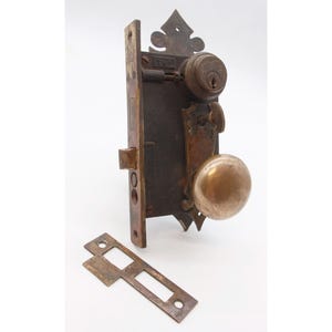 May include: Antique brass door lock with a decorative keyhole cover and a round knob. The lock has a keyhole and a small latch. The knob is round and has a smooth surface. The lock is attached to a metal plate.