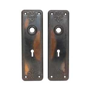 Puede incluir: Un par de placas de puerta antiguas con diseños ornamentados y agujeros de llave. Las placas son de metal y tienen un acabado marrón oscuro.