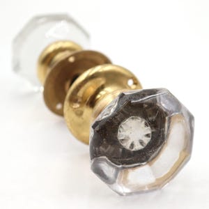 May include: A collection of vintage door knobs. The knobs feature clear, faceted glass and brass hardware. The brass components have a warm, golden hue. The knobs are arranged in a row against a white background.