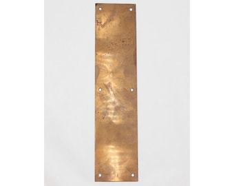 May include: A rectangular brass door plate with a weathered finish. The plate has four evenly spaced holes and a slightly textured surface. The brass has a warm, golden hue, and the plate is vertically oriented.