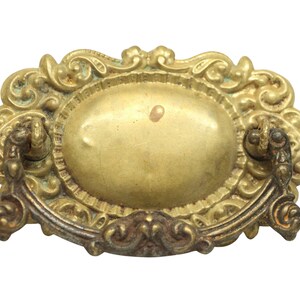 May include: Ornate gold-toned metal drawer pull with a decorative oval centre and a curved handle.