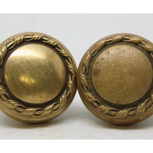 May include: Two antique brass door knobs. Each knob features a circular centre and is encircled by a rope-like design. The brass has a warm, aged patina, suggesting vintage use. The knobs are likely for interior or exterior doors.