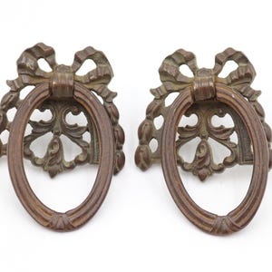 May include: Two antique bronze-colored drawer pulls. Each pull features an oval handle, a decorative bow, and ornate floral detailing. These vintage hardware pieces are ideal for adding a touch of elegance to furniture.