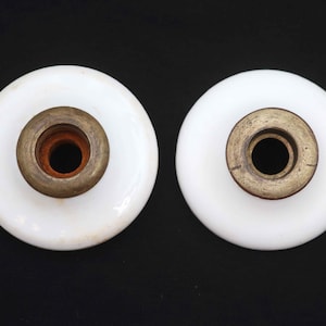 May include: Two white, circular cabinet knobs with brass-colored metal centers. The knobs have a slightly domed shape and are set against a black background. The metal centers have a central hole.
