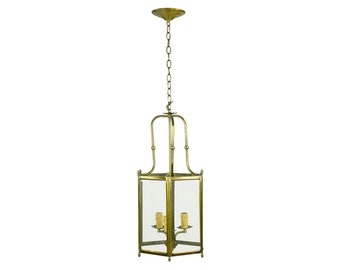 May include: A brass-colored, rectangular pendant light fixture with clear glass panels. The fixture has a decorative top and a chain for hanging. Inside are three candle-style light bulbs.
