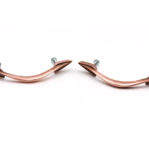 May include: Two copper cabinet pulls with a curved shape and arrowhead ends. The pulls have a brushed finish.