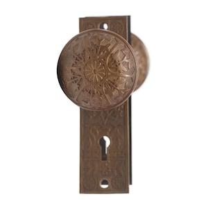 May include: A vintage-style door knob and backplate with an ornate design. The knob and backplate are made of a dark brown metal.