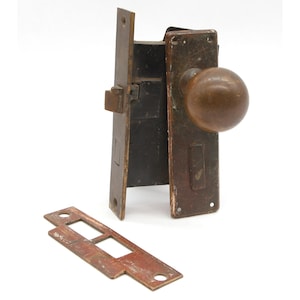 May include: A vintage brass door lock set with a round knob and a separate strike plate. The lock is in a worn, aged condition, with a dark brown patina.
