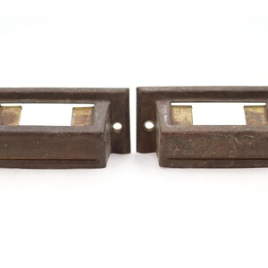 May include: Two rectangular metal drawer pulls with a dark brown, weathered finish. Each pull features a central label holder slot with a rectangular opening and two small circular screw holes on the flat side flanges for mounting to furniture.