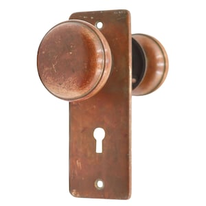 May include: Antique bronze door knob set with a keyhole. The set includes two round knobs and a rectangular backplate. The hardware has a weathered appearance, suggesting age and use. The door hardware is likely for a vintage or historic home.