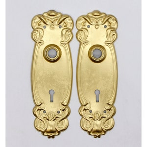 Op de afbeelding: Twee sierlijke, goudkleurige deurplaten met een sleutelgat en een rond gat in het midden. De platen hebben een decoratief ontwerp met krulvormen en bloemmotieven.
