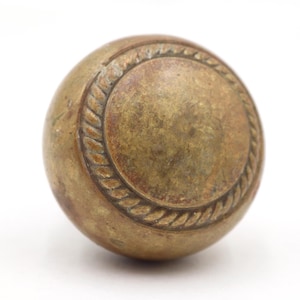 May include: A vintage brass door knob with a round shape. The knob has a central circular design with a rope-like border. The brass has a warm, aged patina, suggesting its antique nature. The knob is set against a white background.