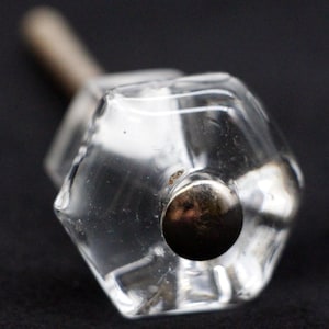 Pode incluir: Um botão de armário de vidro transparente com um centro de metal prateado. O botão tem uma forma hexagonal e é projetado para ser usado em móveis.