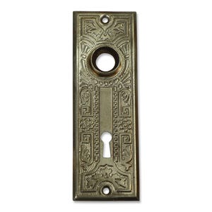 May include: Antique brass-toned door plate with ornate floral and geometric relief patterns. The rectangular plate features a keyhole, a circular opening, and mounting holes. The design is symmetrical and detailed, showcasing a vintage aesthetic.