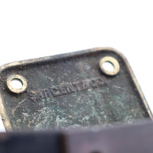 May include: Close-up of a rectangular, aged metal plate with two circular holes and the text "SARGENT & CO." embossed on its surface. The plate has a weathered, greenish-brown patina, suggesting age and use. The background is white.