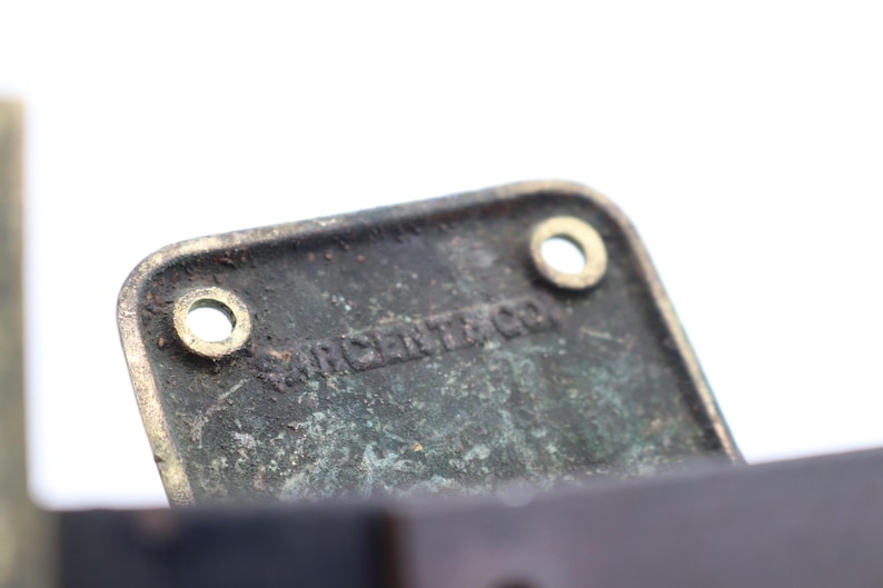 May include: Close-up of a rectangular, aged metal plate with two circular holes and the text "SARGENT & CO." embossed on its surface. The plate has a weathered, greenish-brown patina, suggesting age and use. The background is white.