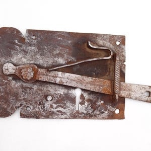 May include: A rusty, antique metal lock with a keyhole and a sliding bolt. The lock has a decorative, floral-shaped element and a curved latch. The metal shows signs of age and wear, with a mottled brown and gray appearance.