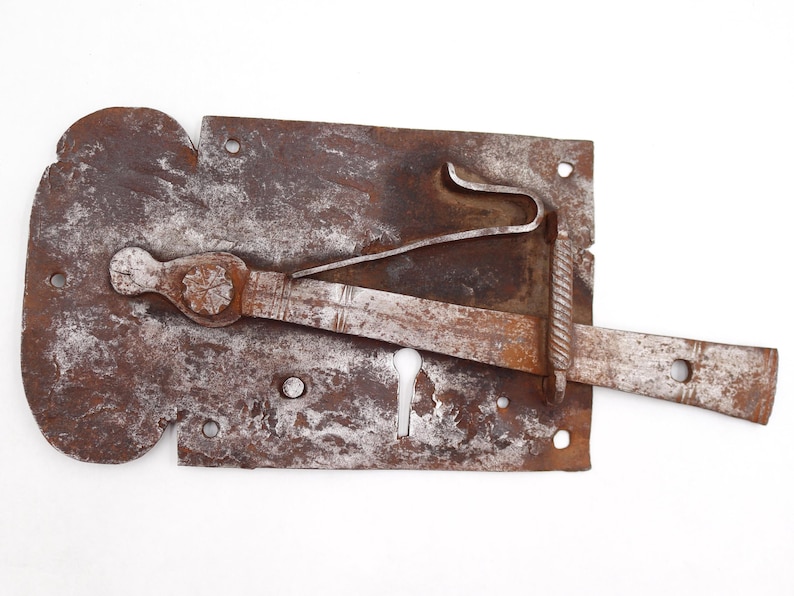 May include: A rusty, antique metal lock with a keyhole and a sliding bolt. The lock has a decorative, floral-shaped element and a curved latch. The metal shows signs of age and wear, with a mottled brown and gray appearance.