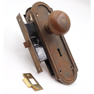 May include: An antique door lock set with a brown, rounded doorknob and a rectangular, ornate backplate. The set includes a latch mechanism and a separate strike plate, all with a weathered, aged appearance.