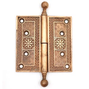 May include: A bronze-colored door hinge with an ornate design. The hinge features a rectangular shape with a woven pattern and decorative sunburst motifs. It has a central barrel and ball finials, suitable for vintage or antique doors.