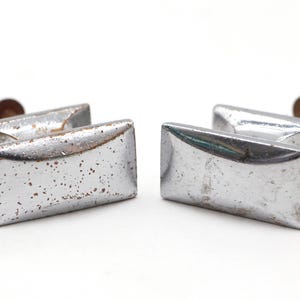 Könnte beinhalten: Vier Vintage-Schubladengriffe aus silberfarbenem Metall mit rechteckiger Form und abgerundeten Kanten. Die Griffe weisen ein leicht abgenutztes Aussehen mit Rostflecken auf. Jeder Griff hat einen kleinen, runden, braun getönten Metallstift.