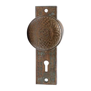 May include: Antique brass door knob with a decorative pattern and a keyhole. The knob is round and has a raised design. The backplate is rectangular and has a similar design.
