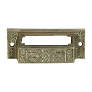 Może przedstawiać: Antyczny mosiężny uchwyt do szuflady z ozdobnym wzorem kwiatowym. Uchwyt ma prostokątny kształt z wycięciem w środku na etykietę.