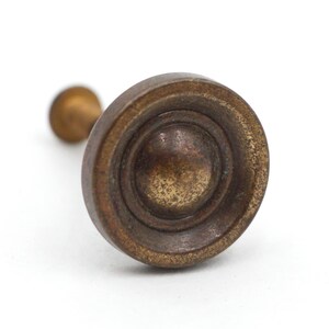 May include: Close-up of a vintage brass drawer pull with a circular design. The pull has a weathered, antique appearance with a central raised circle and concentric rings. The stem is a lighter shade of brass.